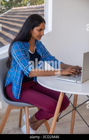 Indische Frau, die auf grauem Stuhl sitzt und auf silbernem Laptop am runden weißen Tisch neben dem Fenster schreibt Stockfoto