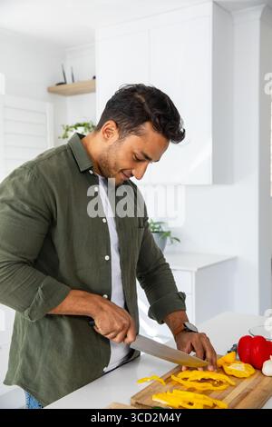 Mann, der ein Messer schwingt, der gelbe Paprika neben rotem Pfeffer auf dem Schneidebrett an der Küchentheke schneidet Stockfoto