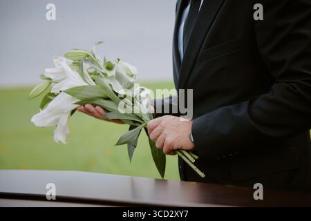 Kaukasier mittleren Alters hält Lilien, die während der Beerdigung im Freien neben dem Sarg stehen, Hände sichtbar, die Blumen in Geste der Erinnerung und des Respekts arrangieren Stockfoto