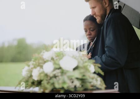 Kaukasischer Mann mittleren Alters, der neben der Schwarzen Frau im mittleren Alter steht und einen Schirm in der Nähe des Sarges mit Blumen während der Begräbniszeremonie im Freien hält Stockfoto