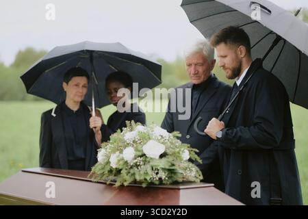 Gruppe von kaukasischen Männern mittleren Alters, älteren kaukasischen Männern, kaukasischen Frauen mittleren Alters, Schwarzen Frauen mittleren Alters, die zusammen unter Regenschirmen stehen und um einen geschlossenen Sarg mit Blumen im Freien trauern Stockfoto