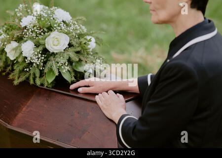 Kaukasische Frau mittleren Alters, die neben einem hölzernen Sarg steht und die Hände auf der Oberfläche liegen, Blumenarrangements mit Rosen und Grün auf der Oberseite, Begräbnisstätte im Freien Stockfoto