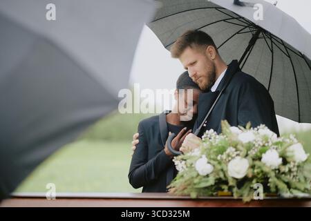 Kaukasischer Mann tröstet Schwarze Frau, während er bei der Beerdigung im Freien unter einem Schirm steht, beide erscheinen emotional und trauern in der Nähe des Sarges mit Blumenarrangements Stockfoto
