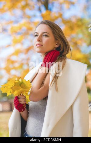 Elegante junge Frau in einem stilvollen weißen Mantel, die tagträumen und einen Haufen bunten, gelben Herbstblätter in der Hand, die in die Ferne starren Stockfoto