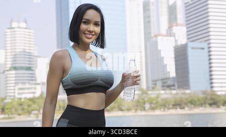 Eine fitte Frau macht während ihrer Trainingsroutine im Park eine Pause und hält eine erfrischende Flasche Wasser mit der Skyline der Stadt im Hintergrund Stockfoto