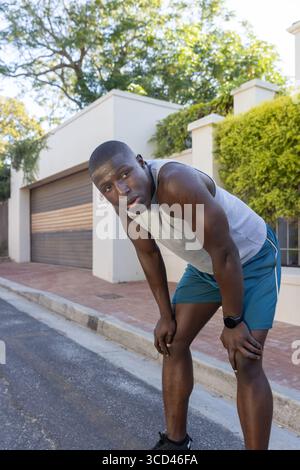 Afroamerikaner, der sich nach vorne beugt und auf der Vorstadtstraße mit Smartwatch und Schuhen Atem schnappt Stockfoto