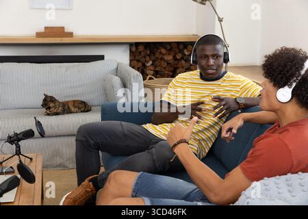 Verschiedene männliche Freunde nehmen Podcasts auf blauem Sofa im Wohnzimmer mit Mikrofonen und Kopfhörern auf Stockfoto