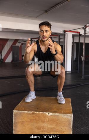 Athletischer Mann hockt auf hölzernem Plyometrikkasten in der Nähe von Kampfseilen und streckt die Arme nach vorne Stockfoto