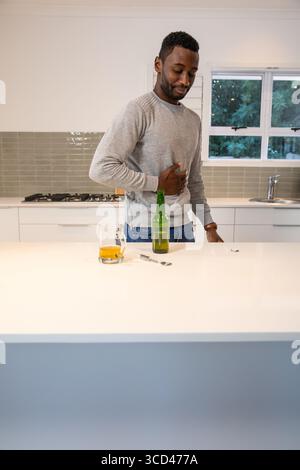 Mittelalter afroamerikanischer Mann, der den Magen drückt und auf der Kücheninsel Bierflasche und Glas ansieht Stockfoto