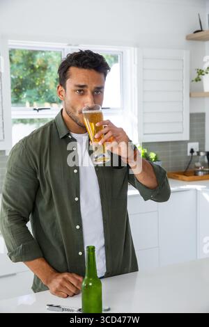 Mann trinkt aus hohem Bierglas, steht auf der weißen Kücheninsel mit grüner Flasche und Korkenzieher Stockfoto