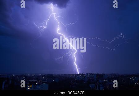 Ein kräftiger Blitz, der nachts durch den dunklen, stürmischen Himmel über der Stadtlandschaft zieht. Die helle Lichtszene des Blitzes bildet einen scharfen Kontrast zu dunklen Wolken und gedämpften Lichtern der darunter liegenden Gebäude. Stockfoto