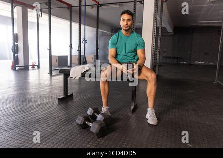 Athletischer Mann, der auf einer Hantelbank im Fitnessraum sitzt und eine digitale Stoppuhr in der Nähe von Kurzhanteln hält Stockfoto