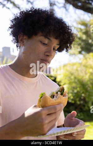 Afroamerikaner mit rosa T-Shirt, der halb gegessen Subsandwich und Papierteller im Garten hält Stockfoto