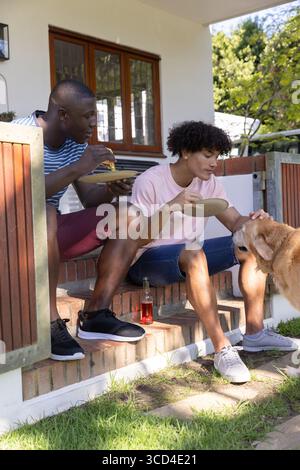 Verschiedene männliche Freunde sitzen auf Steintreppen und essen Pizzascheiben, streicheln Hund, halten Glasflasche Stockfoto