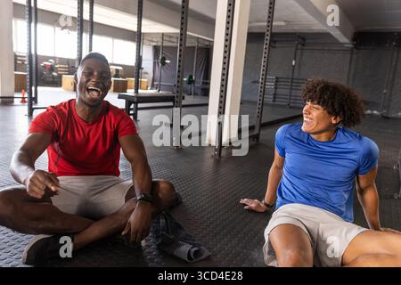 Verschiedene männliche Freunde sitzen in Sportbekleidung auf dem Fitnessraum, lachen und plaudern in der Nähe von Hockregalen Stockfoto