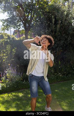 Hispanischer Mann, der auf einem lebhaften Gras im Garten unter einem hohen Baum steht und die Augen vor der Sonne schützt Stockfoto