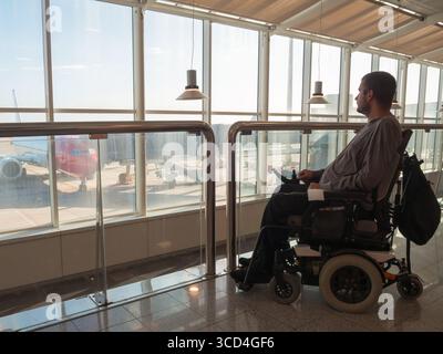 Behinderte Personen, die einen Rollstuhl benutzen, beobachten tagsüber durch große Fenster in einem Flughafenterminal den Rollstuhl und die Flugzeuge. Das helle Stockfoto