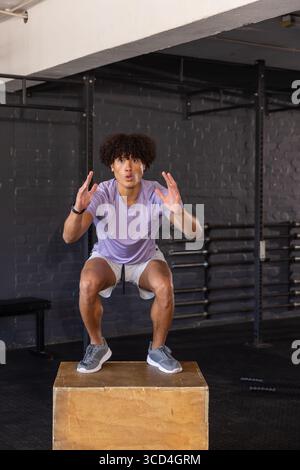 Afroamerikanischer Athlet hockt auf hölzernem Plyometriekasten im Fitnessstudio mit Gewichtsständer Stockfoto