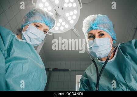 Zwei weibliche Chirurgen in Peelings und Masken konzentrieren sich auf den bevorstehenden Eingriff unter hellem Licht, was ihr Engagement und ihre Teamfähigkeit unterstreicht Stockfoto