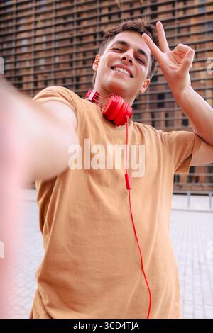 Ein lächelnder junger Mann, der ein Selfie mit einem Friedenszeichen in der Stadt macht, vertikal. Stockfoto