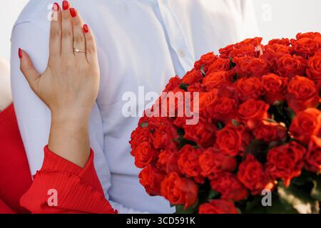 Ein herzerwärmendes Bild zeigt einen Verlobungsvorschlag mit einem funkelnden Diamantring an der Hand einer Frau, der einen großen Blumenstrauß aus leuchtenden roten Rosen hält Stockfoto