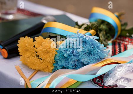 Ein lebhafter Blumenstrauß mit gelben und blauen Chrysanthemen mit Bändern liegt auf einem Tisch, der mit Elementen der ukrainischen Flagge und einem Mikrofon und einer Symbe verziert ist Stockfoto