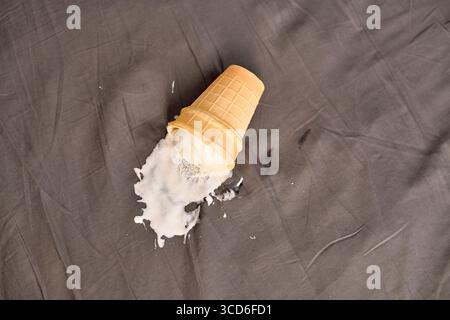 Gefallene Eiskegel verschüttet Inhalt auf dunkles Gewebe. Vanille- und Schokoladeneis sind teilweise geschmolzen, wodurch ein struktureller Kontrast entsteht. Weiches Licht hoch Stockfoto