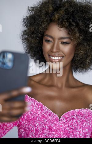 Positive afroamerikanische Frau mit lockigem Haar, die lächelt und in die Kamera schaut, während sie ein Selfie mit dem Handy macht Stockfoto