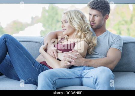 Ein junges Paar, das einen ruhigen Moment zu Hause genießt und sich in den Armen auf einem Sofa unter einem Fenster entspannt Stockfoto