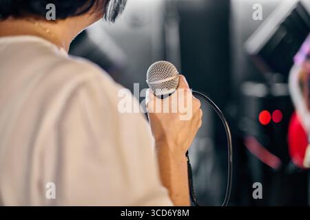 Junge Frau mit schwarzem Haarmikrofon während der lebhaften Bandprobe. Schwach beleuchteter Raum mit konzentrierter, leidenschaftlicher Atmosphäre. Abstrakter Hintergrund Stockfoto