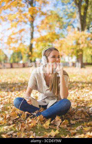 Junge Frau, die sich entspannt beim Hören von Musik auf ihren Kopfhörern entspannt, während sie mit ihrem MP3-Player im Kreuz auf dem Boden sitzt Stockfoto