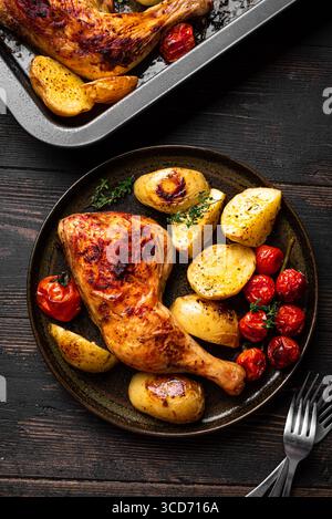 Im Ofen gebratenes Hähnchenschenkelviertel mit Kartoffeln, Tomaten und Kräutern, serviert auf Holztisch, Blick von oben. Herzhafte hausgemachte Mahlzeit - Hühnchen und Gemüse. Stockfoto