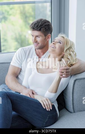 Junges Paar, das einen entspannenden Tag zu Hause verbringt und sich auf einem Sofa unter einem Fenster mit Blick auf den Garten mit einem Lächeln auf die Seite lehnt Stockfoto