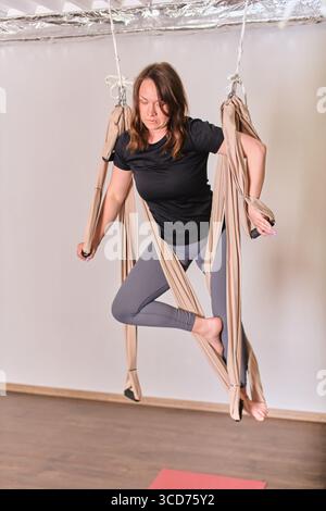 Junge Frau mit braunem Haar übt Luft-Yoga in beiger Hängematte drinnen. Mit schwarzem Hemd und grauer Leggings balanciert er anmutig aus. Hell, ruhig Stockfoto