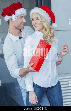 Ein verspielter Mann, der seiner Frau ein Weihnachtsgeschenk schenkt, überrascht sie, als er von hinten in ihren bunten roten Weihnachtsmützen umhergreift Stockfoto