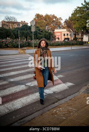 Attraktive Frau in braunem Mantel und Jeans, die auf einer Fußgängerüberquerung läuft und eine alte Filmkamera hält. Ein attraktives Mädchen, das ihren Urlaub genießt und reist Stockfoto
