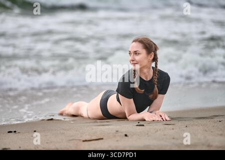 Junge Frau mit braunen Haaren übt Yoga am ostseestrand. Der bewölkte Himmel und sanfte Wellen sorgen für eine ruhige Szene und verstärken den ruhigen Moment. Sandiger Sho Stockfoto