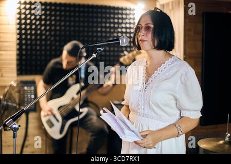 Junge Frau mit schwarzem Haar und Brille hält Noten und singt leidenschaftlich ins Mikrofon. Der Erwachsene Gitarrist konzentriert sich auf den Hintergrund. Warmes Licht Stockfoto