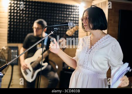 Junge Frau mit schwarzem Haar und Brille hält Noten und singt leidenschaftlich ins Mikrofon. Der Erwachsene Gitarrist konzentriert sich auf den Hintergrund. Warmes Licht Stockfoto