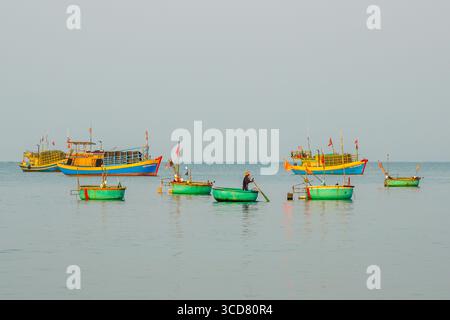 Mui ne, Vietnam, 22. März 2025: Ein einsamer Fischer rudert ein grünes Korbboot in der Nähe größerer Fischereischiffe in den ruhigen Morgengewässern von Mui ne Stockfoto