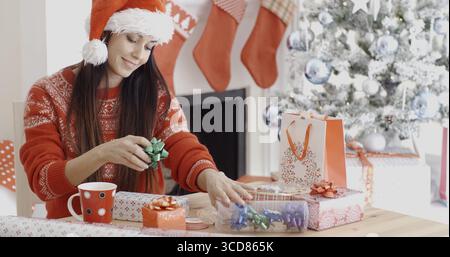 Junge Frau, die ihre Weihnachtsgeschenke mit Schleifen und Bändern dekoriert, während sie an einem Tisch vor einem geschmückten Weihnachtsbaum sitzt Stockfoto
