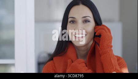 Lächelnde, wunderschöne junge brünette Frau in Wintermode, die zuhause drinnen steht und mit einem freundlichen, glücklichen Lächeln in die Kamera blickt Stockfoto