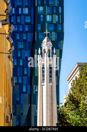 Ein moderner Kirchturm mit Kreuz steht vor dem Hintergrund eines zeitgenössischen Glasgebäudes. Die Szene umfasst kontrastierende architektonische Stile und Stockfoto