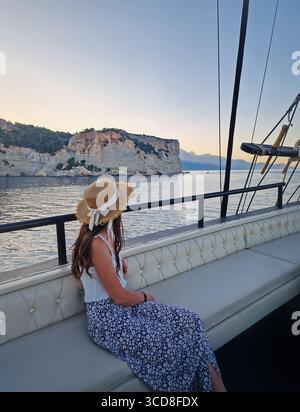 Entspannte junge Touristenfrau mit Strohhut, segelt in einem Segelboot und blickt auf das Meer und die Berge im Hintergrund unter einem Sonnenuntergang. Stockfoto