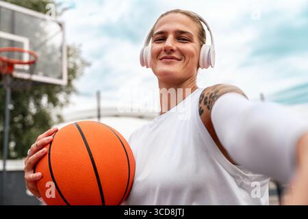 Junge Frau mit Kopfhörern, die Basketball hält und Selfie auf dem städtischen Streetballfeld macht Stockfoto