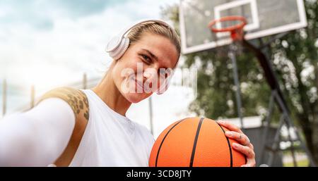 Selbstporträt einer jungen, aktiven Stadtfrau mit Kopfhörern und Basketballhaltung. Stockfoto
