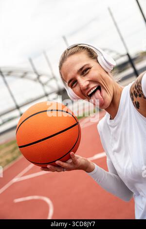 Cooles Attitüdenporträt einer jungen Frau mit Kopfhörern, die Basketball auf dem städtischen Sportplatz hält Stockfoto