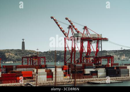 Geschäftiger Containerhafen mit hohen roten Kränen und gestapelten bunten Schiffscontainern vor dem Hintergrund einer Hängebrücke und der Cristo Stockfoto