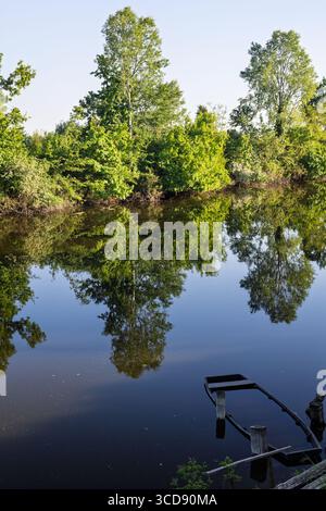 Ruhige Landschaft mit einem ruhigen Fluss und lebendigen grünen Bäumen, die sich auf der spiegelähnlichen Oberfläche des Wassers spiegeln. Ein altes hölzernes Boot verleiht dem einen rustikalen Charme Stockfoto