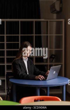 Geschäftsfrau im formellen schwarzen Anzug, die in einem gemütlichen, modernen Büro mit farbenfrohen Stühlen am Laptop arbeitet und sich in die Arbeit hineinsetzt Stockfoto